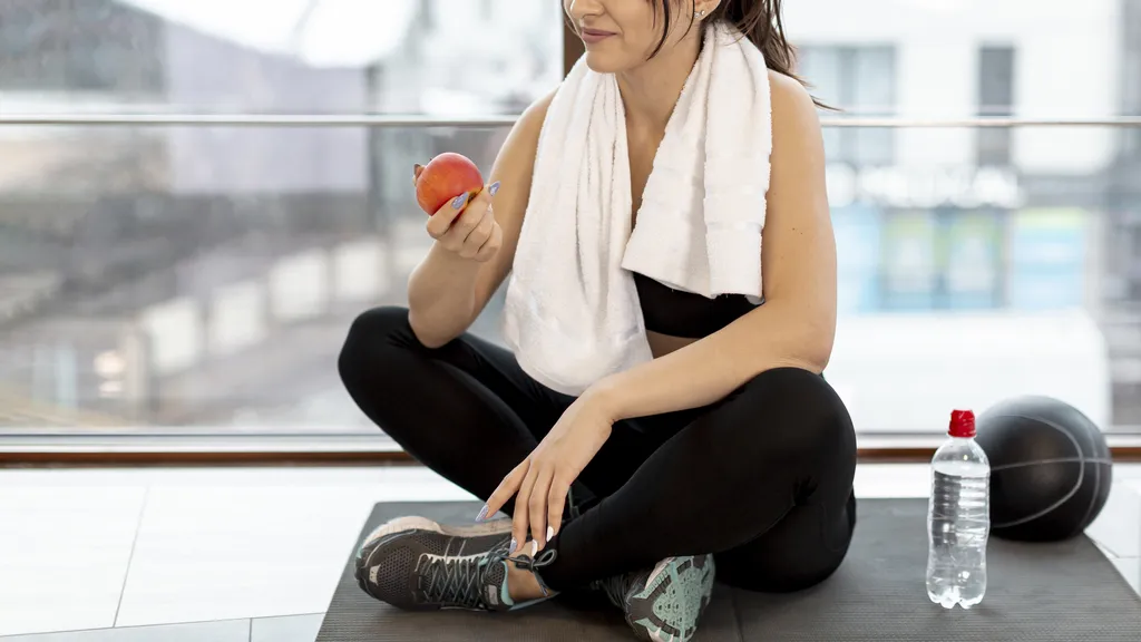 Woman in sportswear sitting on a gym mat, eating an apple during a workout break, with a towel around her neck, a water bottle, and a medicine ball nearby.